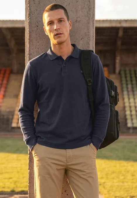 Langarm Poloshirt in Waffelstruktur dark ink blue Ein Mann mit Rucksack steht vor Stadiontribüne, trägt dunkelblaues Polo und helle Hose.