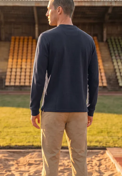 Langarm Poloshirt in Waffelstruktur dark ink blue Ein Mann in blauem Langarmshirt und beiger Hose steht von hinten vor Tribünen auf einer Sportanlage.