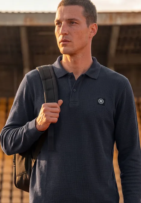 Langarm Poloshirt in Waffelstruktur dark ink blue Mann mit Rucksack und dunkelblauem Poloshirt steht vor einer überdachten Bahnhofskulisse.