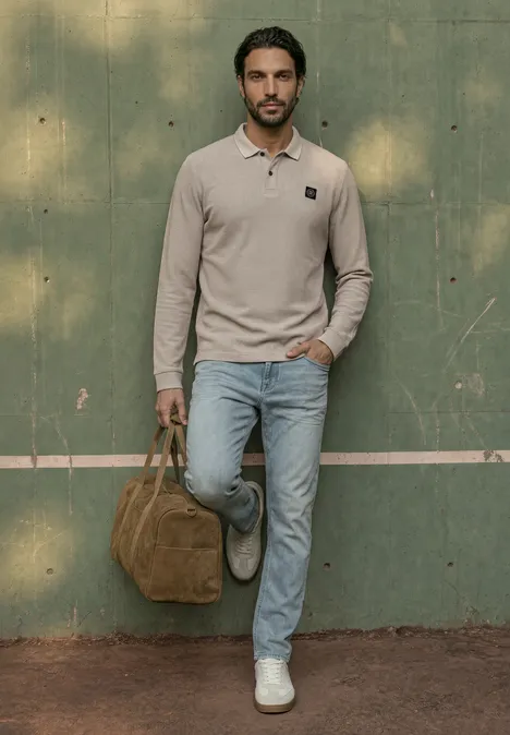 Langarm Poloshirt in Waffelstruktur antique beige Ein Mann lehnt an einer grünen Wand, trägt ein beiges Polo, Jeans, weiße Sneaker und hält eine braune Tasche.