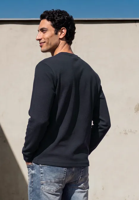 Ein junger Mann steht seitlich, trägt schwarzen Pullover und Jeans vor einer beigen Wand.