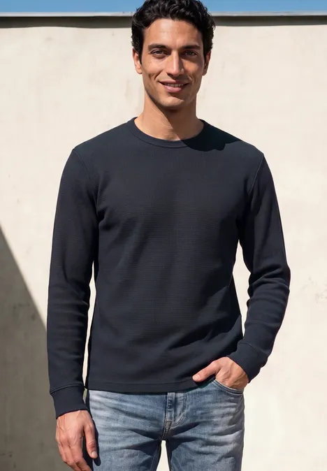 Ein lächelnder Mann in schwarzem Pullover und Jeans steht vor heller Wand im Freien.