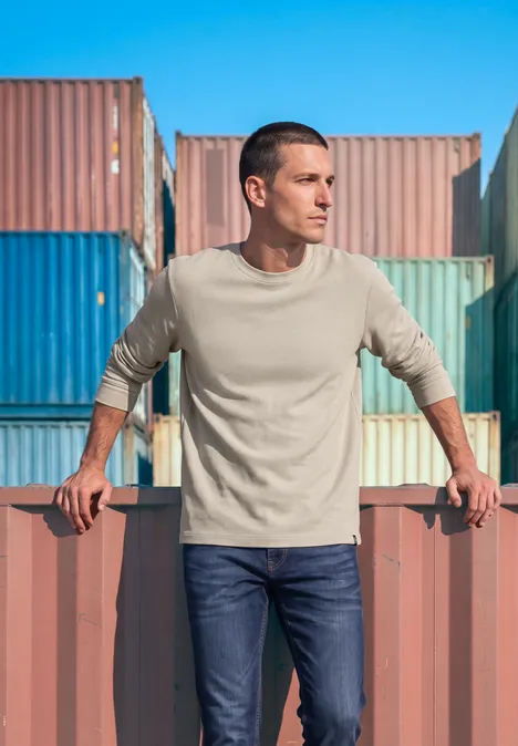 Ein Mann lehnt an bunten Frachtcontainern im Hafen, trägt beige Pullover und Jeans.