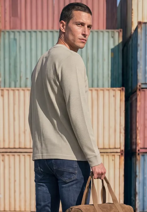 Ein Mann in beige Pullover und Jeans, hält eine Tasche und steht vor bunten Containerwänden.