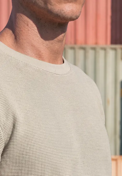 Ein Mann trägt ein beiges Waffelstrickshirt vor bunten Containerwänden.