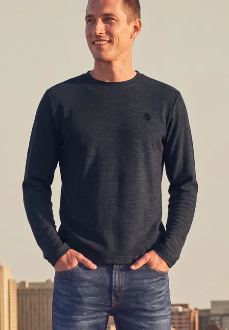 Un homme souriant en pull gris foncé et jean, mains dans les poches, devant la skyline urbaine au coucher du soleil.