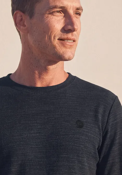 Homme en tee-shirt manches longues gris foncé, regard latéral, lumière chaude, logo discret sur la poitrine.