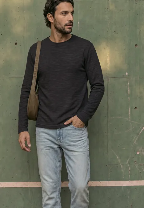 Langarm Shirt mit Rundhals in Unifarbe basic black Ein Mann mit dunkelgrauem Pullover, Jeanshose und Umhängetasche steht vor einer grünen Wand.