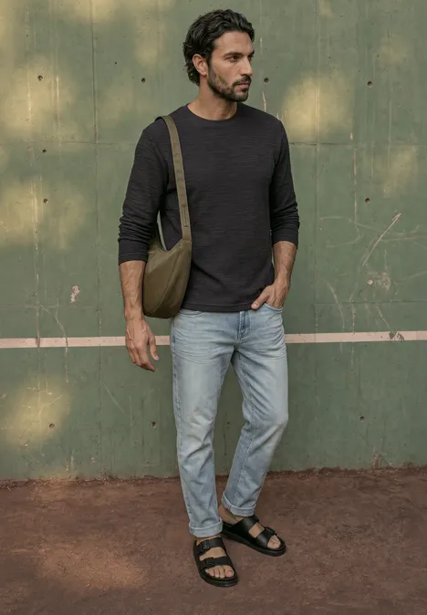 Langarm Shirt mit Rundhals in Unifarbe basic black Ein Mann in schwarzem Langarmshirt, hellen Jeans und Sandalen steht vor grünem Hintergrund mit Umhängetasche.