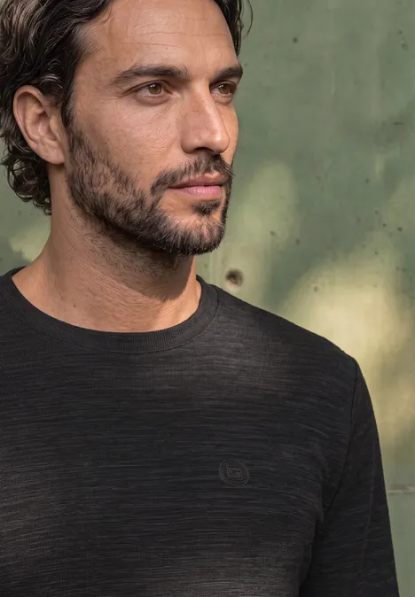 Langarm Shirt mit Rundhals in Unifarbe basic black Porträt eines nachdenklichen Mannes mit dunklem Haar und Bart vor unscharf grünem Hintergrund.