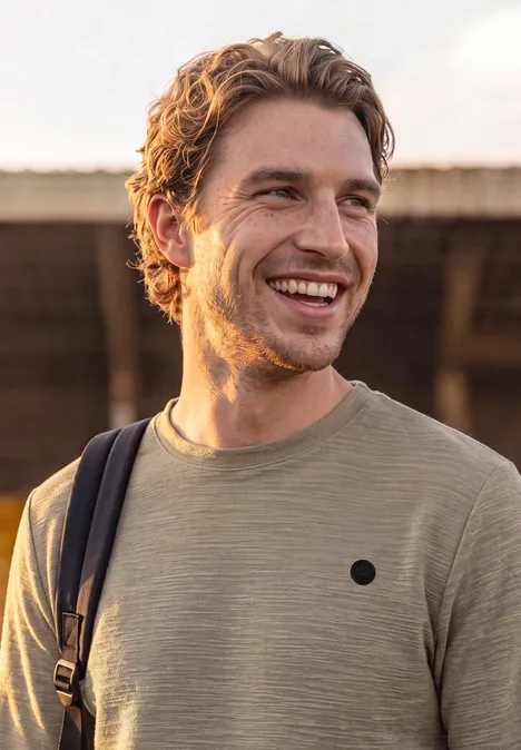 Langarm Shirt mit Rundhals in Unifarbe antique beige Ein junger Mann in einem beigen T-Shirt mit Rucksackschultergurt lächelt im Freien.