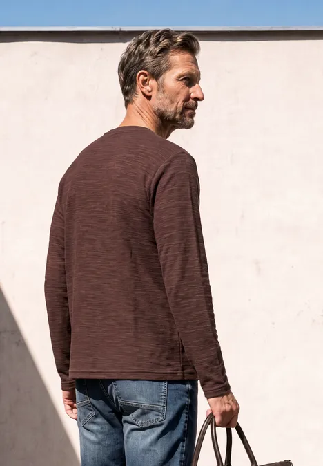 Langarm Shirt mit Rundhals in Unifarbe maroon brown Ein Mann steht seitlich mit braunem Langarmshirt, Jeans und hält eine Tasche in der Hand vor heller Wand.