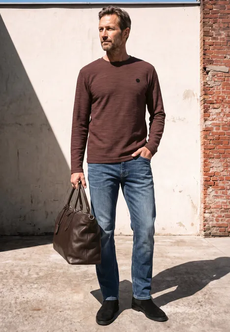 Langarm Shirt mit Rundhals in Unifarbe maroon brown Mann im braunen Pullover, blauen Jeans und schwarzen Schuhen trägt eine große Ledertasche, steht im Sonnenlicht.