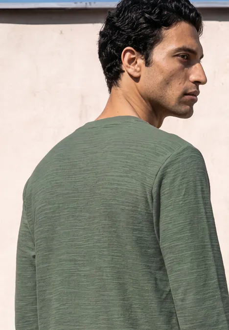 Langarm Shirt mit Rundhals in Unifarbe willow green Ein Mann steht im Profil, trägt ein grünes Langarmshirt und blickt seitlich vor einer hellen Wand.