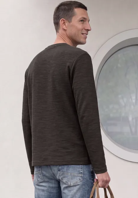Ein Mann steht von hinten vor einem runden Fenster, trägt dunkelgraues Oberteil und Jeans und hält eine Tasche.