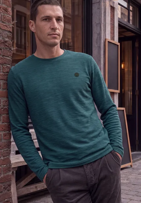 Un homme appuyé sur un mur de briques, vêtu d’un t-shirt à manches longues vert et d’un pantalon gris.