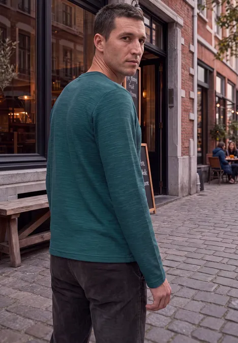 Homme en pull vert debout sur pavés, regard par-dessus l'épaule.