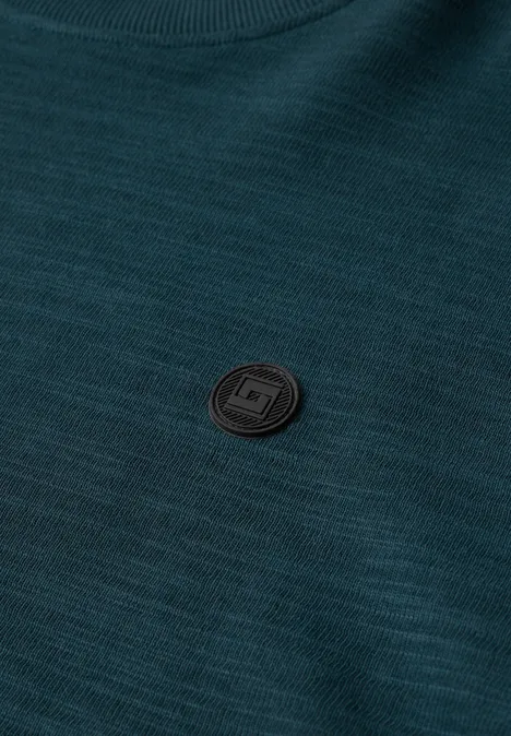 Un pull vert foncé à texture fine, avec un petit écusson rond noir sur la poitrine.