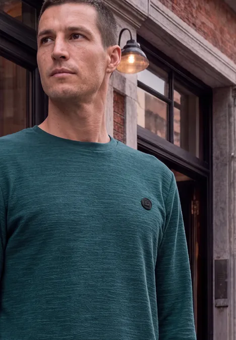 Homme en sweat vert à manches longues avec logo rond devant une façade en brique.