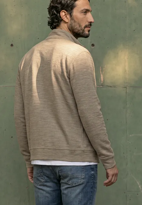 Ein Mann steht mit dem Rücken zur Kamera, trägt beiges Cardigan, weißes T-Shirt und Jeans vor grüner Wand.
