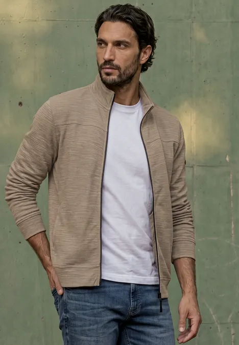 Ein Mann in beiger Strickjacke, weißem T-Shirt und Jeans vor grünem Hintergrund.