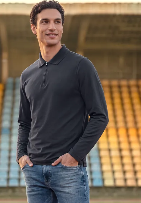 Ein lächelnder Mann in schwarzem Langarmpoloshirt und Jeans steht vor leeren Stadionrängen.