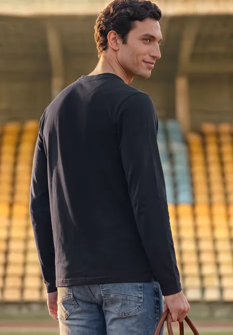 Ein Mann im schwarzen Langarmshirt und Jeans blickt lächelnd über die Schulter in einem Stadion.