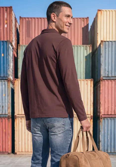 Ein Mann trägt ein dunkelrotes Poloshirt, Jeans und eine Tasche vor bunten Frachtcontainern.