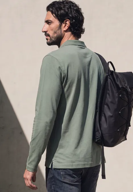 Ein Mann mit grünem Langarmshirt und Rucksack schaut seitlich vor einer hellen Wand.