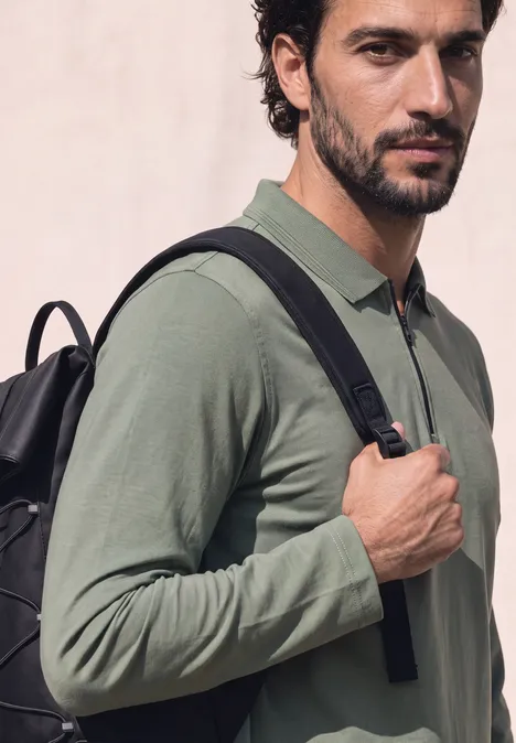 Ein Mann mit dunklen Haaren trägt einen Rucksack und ein langärmeliges, khakifarbenes Poloshirt.