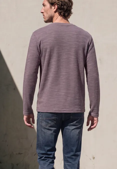 Ein Mann von hinten trägt ein langärmeliges violettes Shirt und Jeans vor einer grauen Wand mit Schatten.
