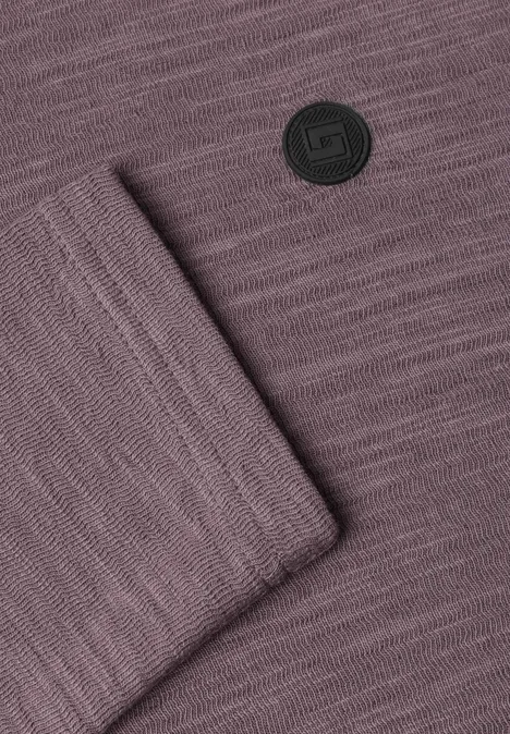 Violetter Strickstoff mit sichtbarer Maschenstruktur, umgeschlagener Ecke und rundem schwarzen Logo.
