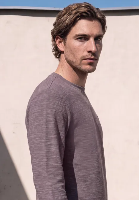 Ein Mann mit braunen Haaren trägt einen grauen Pullover vor einer hellen Mauer.