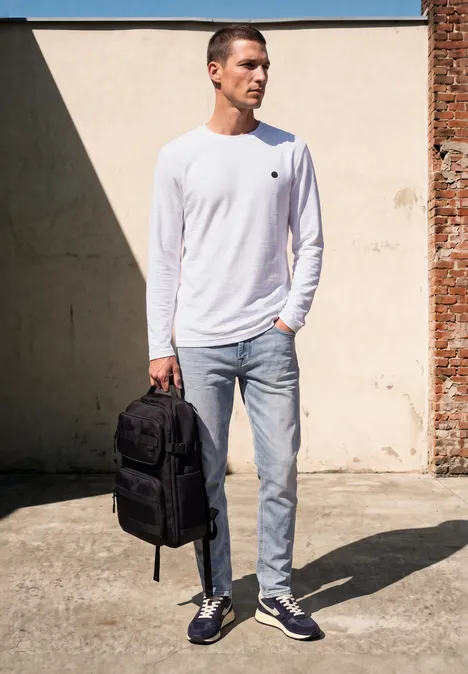 Ein Mann in weißem Langarmshirt und Jeans trägt einen schwarzen Rucksack und steht vor einer hellen Mauer.