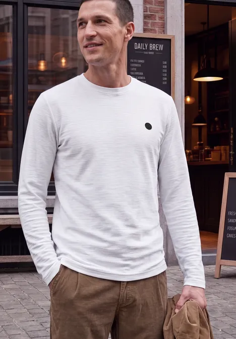 Langarm Shirt mit Rundhals in Unifarbe bright white Ein Mann in weißem Langarmshirt und brauner Hose steht vor einem Café und lächelt leicht.