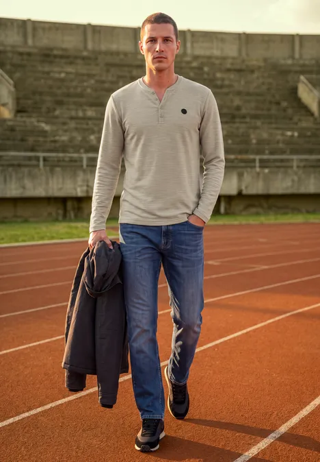 Un homme en jean et t-shirt se tient sur une piste d'athlétisme en tenant une veste.