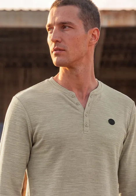 Homme en t-shirt Henley vert clair regarde au loin devant un décor rustique