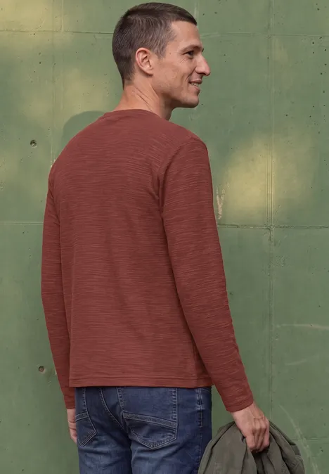 Un homme vu de côté porte un t-shirt à manches longues rouge-brun, un jean et tient une veste à la main.