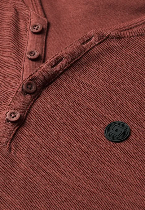 Gros plan d'un t-shirt Henley rouille avec patte de boutonnage et patch logo rond noir.