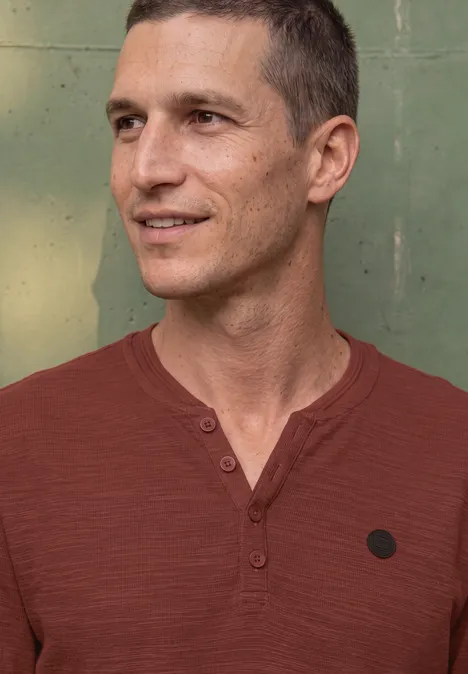 Un homme souriant portant un t-shirt rouge devant un mur vert.