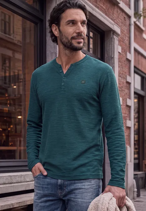 Un homme en t-shirt vert à manches longues et jean pose décontracté dans la rue.
