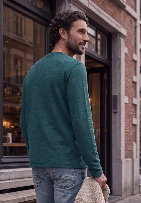 Un homme en t-shirt vert à manches longues et jean marche devant un bâtiment tout en regardant par-dessus son épaule.