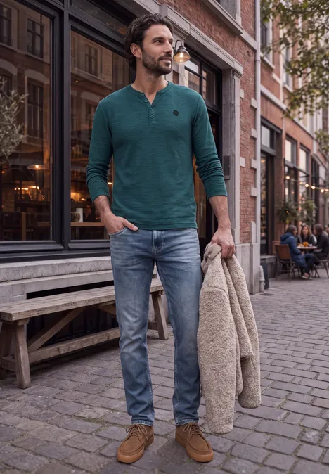 Un homme élégant en pull vert et jean se promène tranquillement dans une ruelle urbaine, manteau à la main.