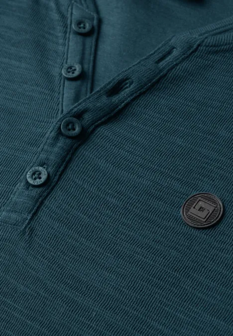Gros plan sur un pull côtelé bleu pétrole avec patte de boutonnage et écusson rond.