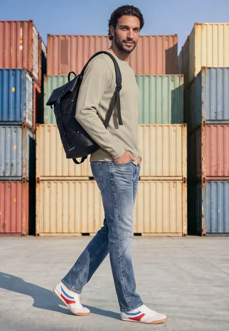 Un homme en jean, baskets blanches et sac à dos se tient de profil devant des conteneurs colorés.