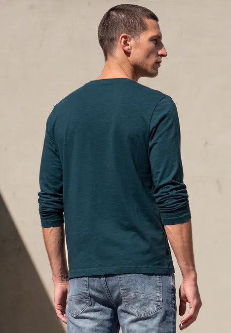 Mann steht mit dem Rücken zur Kamera, trägt grünes Langarmshirt und Jeans vor heller Wand im Sonnenlicht.