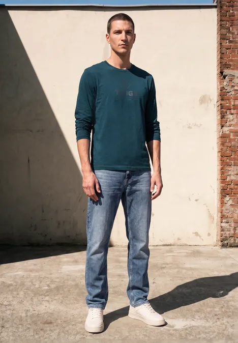 Ein Mann in einem grünen Pullover und Jeans steht selbstbewusst vor einer Wand und schaut in die Kamera.