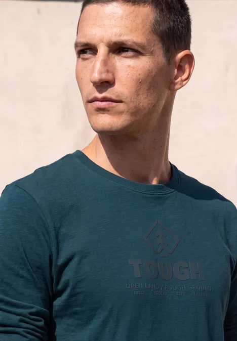 Mann mit kurzem Haar in grünem T-Shirt mit 'TOUGH'-Schriftzug blickt nach rechts.