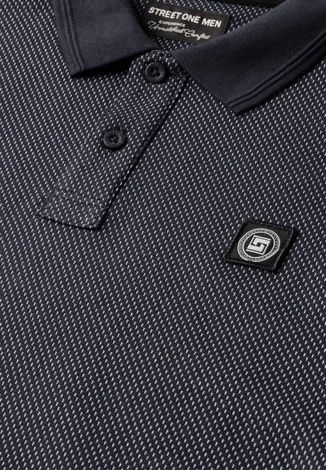 Gros plan sur un polo noir à motif discret, col et épingle siglée S1 bien visible.
