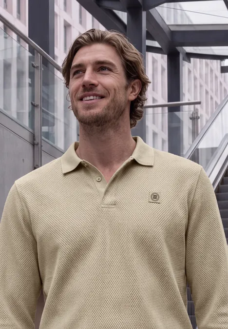 Un homme souriant en polo beige devant une façade en verre moderne.